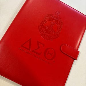May include: A red leather binder with the Greek letters Delta Sigma Theta and the organization's crest embossed on the front.