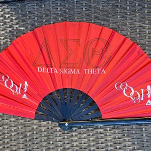 Puede incluir: Un abanico plegable rojo con detalles en negro. El abanico presenta las letras griegas Delta, Sigma y Theta, junto con las palabras "Delta Sigma Theta". El abanico también tiene la palabra "COOP!" y el año "1913" impresos.