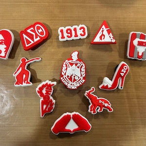 May include: A collection of red and white shoe charms with various designs, including a heart with a face, Greek letters, the number 1913, a high heel, and an elephant. The charms are on a brown surface.