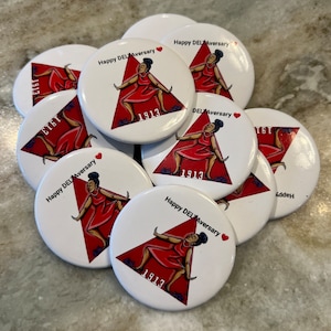 May include: A stack of white round buttons with a red triangle and a black woman in a red dress. The buttons have the text "Happy DEL Aversary" and "1913" on them.