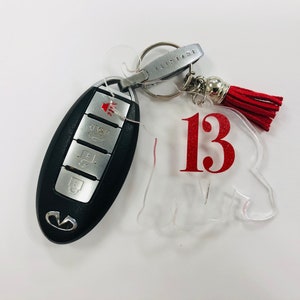 May include: A clear acrylic keychain with the number 13 in red lettering. The keychain is attached to a silver key ring with a red tassel.