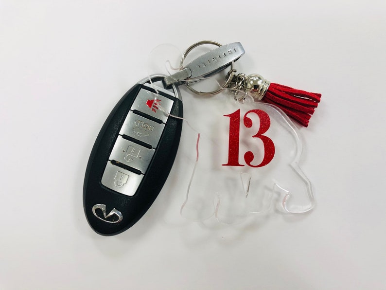 May include: A clear acrylic keychain with the number 13 in red lettering. The keychain is attached to a silver key ring with a red tassel.