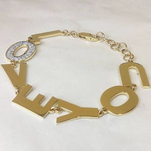 Puede incluir: Pulsera de oro con la palabra "LOVE" escrita en letras individuales. La "O" est&aacute; adornada con peque&ntilde;as piedras transparentes.