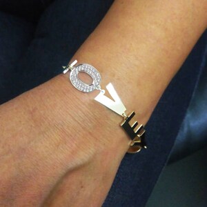 Puede incluir: Pulsera de oro con la palabra "LOVE" escrita en letras grandes. La "O" est&aacute; adornada con diamantes de imitaci&oacute;n transparentes.