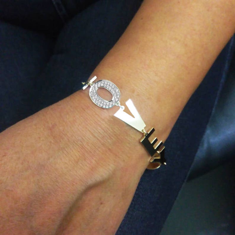 Puede incluir: Pulsera de oro con la palabra "LOVE" escrita en letras grandes. La "O" est&aacute; adornada con diamantes de imitaci&oacute;n transparentes.