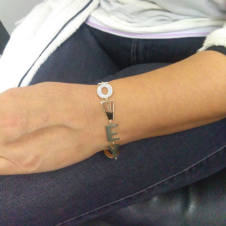 Puede incluir: Pulsera de oro con la palabra "LOVE" escrita en letras grandes. Cada letra est&aacute; decorada con peque&ntilde;as piedras transparentes.