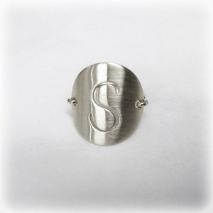 以下が含まれることがあります： 楕円形の顔を持つ銀色のリング。顔の中央には文字「S」が切り抜かれています。リングはつや消し金属仕上げで、両側に小さな装飾的なアクセントが付いています。