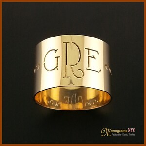 May include: A gold napkin ring with the letters "GRE" cut out. The ring has a polished finish and a simple, elegant design. The bottom of the ring has etched text. The background is black.