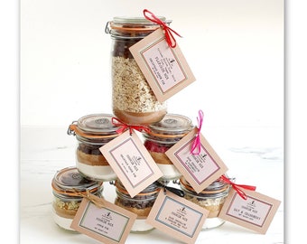 May include: Six glass jars filled with cookie mix ingredients, each with a different flavour. The jars are stacked in a pyramid shape and have brown paper tags with the flavour names: Apple Pie, Double Chocolate & Sea Salt, Oat & Cranberry, Pumpkin Spice, Chocolate Chip, and Snickerdoodle.