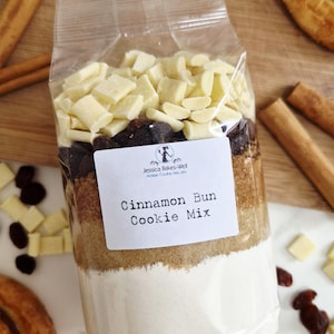 May include: A clear plastic bag containing layered dry ingredients for a Cinnamon Bun Cookie Mix, topped with white chocolate chips and raisins. A white label features the text Jessica Bakes-Well Artisan Cookie Mixes and Cinnamon Bun Cookie Mix.