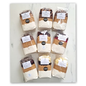 May include: Nine clear plastic bags filled with cookie mix ingredients. Each bag has a label with the name of the cookie flavour and the words "Great Taste". The flavours are: Chocolate Salted Caramel, Sticky Toffee Pudding, Galaxy, Terry's Chocolate Orange Cookie, Apple Pie, Dark Chocolate & Raspberry, Dark Chocolate & Cranberry, White Chocolate & Cranberry, Terry's White Chocolate Orange Cookie.