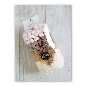 May include: A clear plastic bag filled with ingredients for a s'mores cookie mix. The bag contains a white cookie mix, brown chocolate chips, and pink and white marshmallows. The bag has a label that says "S'mores Cookie Mix" and a "Great Taste" sticker.