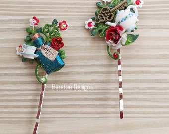 Op de afbeelding: Twee Alice in Wonderland-haarclips. Eén met een blauwe fles met de woorden "Drink Me" en een theekopje. De andere met een witte theepot met een sleutel en een rode roos.