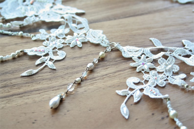 May include: White lace choker necklace with pearl and crystal accents. The necklace has a delicate floral design and features a dangling pearl drop.