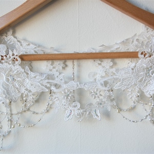May include: A white lace bridal sash with beaded fringe. The sash is hanging on a wooden hanger.