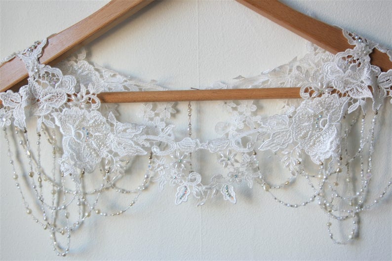May include: A white lace bridal sash with beaded fringe. The sash is hanging on a wooden hanger.