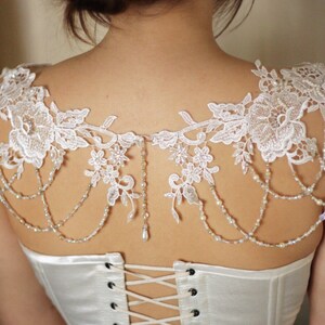 May include: White lace bridal shrug with beaded straps and a delicate floral pattern. The shrug is worn over a white satin corset with a lace-up back.