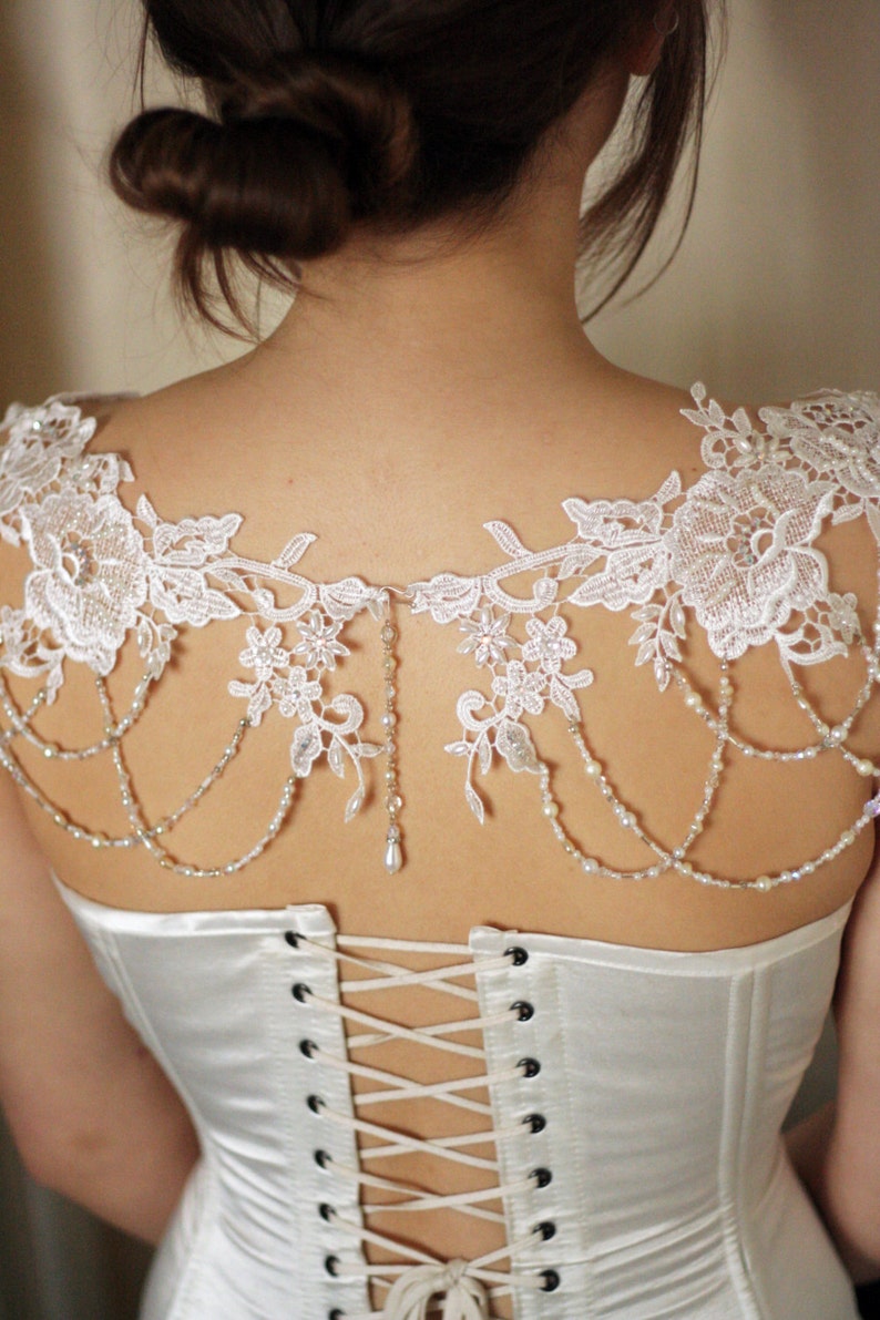 May include: White lace bridal shrug with beaded straps and a delicate floral pattern. The shrug is worn over a white satin corset with a lace-up back.