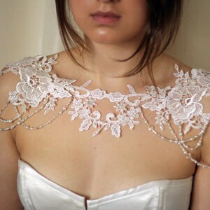 May include: White lace bridal shoulder piece with pearl beading. The lace has a floral design and the beads are strung in a delicate chain.
