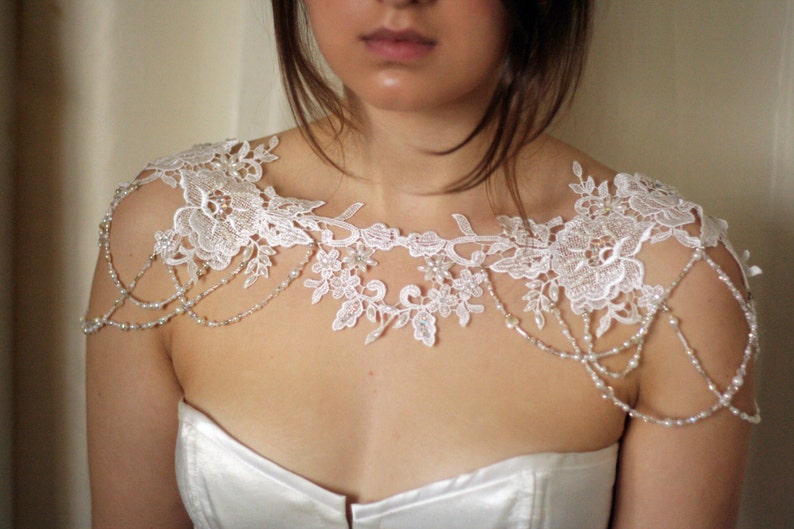 May include: White lace bridal shoulder piece with pearl beading. The lace has a floral design and the beads are strung in a delicate chain.