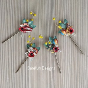 May include: Four handmade hair clips with a whimsical circus theme. Each clip features a red poppy flower, green leaves, and various circus-themed charms, including a rainbow, a carousel horse, a hot air balloon, and a Ferris wheel. The clips are adorned with yellow beads and crystals.