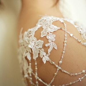 May include: White lace shoulder piece with beaded straps. The lace is delicate and floral, and the straps are made of small pearls and crystals.
