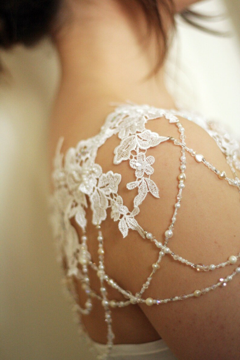 May include: White lace shoulder piece with beaded straps. The lace is delicate and floral, and the straps are made of small pearls and crystals.