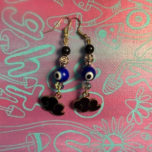 May include: A pair of gold-toned dangle earrings. Each earring features a black bead, a silver bead, a blue evil eye bead, and a clear crystal bead. A black cloud charm hangs from the bottom of each earring.