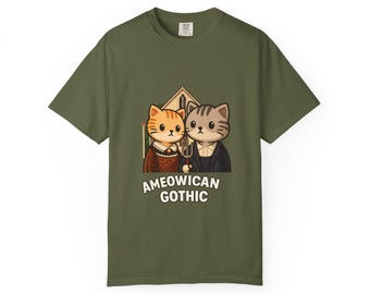 May include: An olive green t-shirt with a cartoon illustration of two cats, styled after the American Gothic painting. The image includes a church and the text "AMEOWICAN GOTHIC" below the cats.