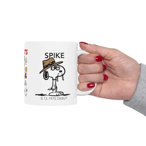 Può includere: Tazza in ceramica bianca con un'illustrazione a cartoni animati di Spike dei Peanuts con un cappello marrone. La parola "SPIKE" è stampata sopra il personaggio, con "8.13.1975 DEBUT" sotto. La tazza ha un manico bianco e una piccola illustrazione laterale.