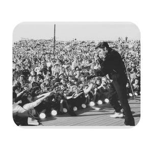 May include: A black and white photo of a musician performing on stage in front of a large crowd. The musician is wearing a dark suit and is holding a microphone. The crowd is reaching out to the musician with their hands.