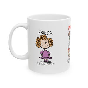 May include: White ceramic mug featuring Frieda from Peanuts, with her name and debut date. The illustration shows Frieda with curly orange hair, wearing a purple dress. The mug has a large handle for easy gripping.