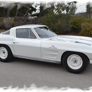 May include: A white 1963 Chevrolet Corvette Stingray sports car with black racing stripes. The car is parked on a paved surface with trees and bushes in the background.