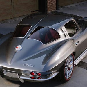 May include: A silver 1963 Chevrolet Corvette Stingray coupe with red taillights and a chrome Corvette emblem. The car is parked on a gray concrete surface.