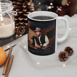 May include: White ceramic coffee mug featuring a vintage image of a man in a cowboy hat and vest. The mug has a black rim and a white handle. The mug is surrounded by decorative items, including a candle, pine cones, and cinnamon sticks.