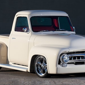 Könnte beinhalten: Ein klassischer weißer Ford Pickup aus den 1950er Jahren mit Chrom-Akzenten und rotem Innenraum. Der LKW steht auf einer grauen Asphaltfläche.