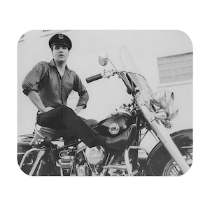 May include: Black and white photograph of a person sitting on a vintage Harley-Davidson motorcycle. The person is wearing a dark cap, a polka-dot shirt, and black pants. The motorcycle has a windshield and chrome details.