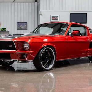 May include: A bright red 1967 Ford Mustang with black accents and black wheels. The car is in a garage setting with a white tiled wall in the background.