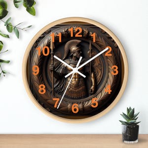 Puede incluir: Un reloj de pared redondo con marco de madera clara. La esfera del reloj presenta un relieve de bronce de un guerrero griego sosteniendo una lanza. Los números en naranja marcan las horas y las manecillas blancas indican la hora.