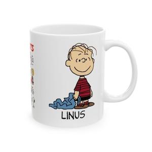 May include: White ceramic mug featuring a cartoon illustration of Linus from the Peanuts comic strip. Linus is depicted in a red and black striped shirt, holding a blue blanket. The mug also has text from the comic strip.