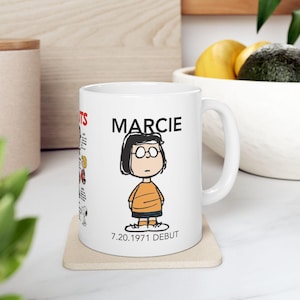 May include: White ceramic mug featuring a cartoon illustration of Marcie from Peanuts. The mug has the name "MARCIE" above the character and the text "7.20.1971 DEBUT" below. The mug is on a beige coaster.