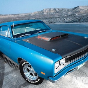May include: A blue 1969 Dodge Super Bee muscle car with a black hood and a scoop. The car is parked on a paved surface with a mountain range in the background.