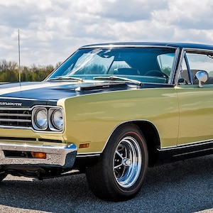 May include: A yellow and black 1969 Plymouth Road Runner with a black vinyl roof and black racing stripes. The car has a chrome bumper and chrome wheels.