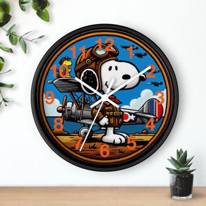May include: A round wall clock with a black frame. The clock face features Snoopy as a World War I flying ace in a brown leather helmet and jacket, with a biplane and blue sky background. Orange numerals mark the hours.