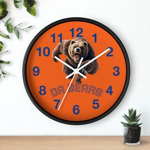 May include: Round wall clock with a black frame and an orange face. The clock features blue numerals and the words "DA BEARS". A brown bear graphic is in the center.