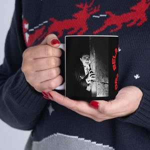 Könnte beinhalten: Eine schwarze Kaffeetasse mit einem Schwarz-Weiß-Bild von Bela Lugosi als Dracula. Auf der Tasse steht der Text "DR. BELA" in Rot. Die Tasse wird von einer Person gehalten, die einen marineblauen Pullover mit einem rot-weißen Rentier-Muster trägt.
