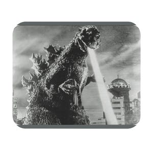 May include: Black and white image of Godzilla, a giant monster, emitting a beam of energy. The image is a still from a classic film, with a cityscape in the background. The image is in a rectangular shape.