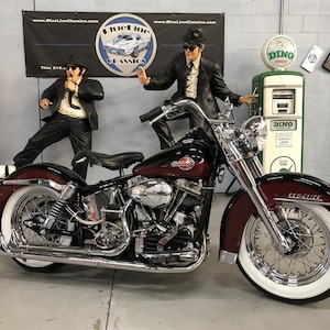 May include: A black and red Harley Davidson motorcycle with whitewall tires and chrome accents. The motorcycle has a black leather seat and a chrome exhaust pipe. The motorcycle is parked in a garage with a gas pump and a sign that says "Blue Line Classics".