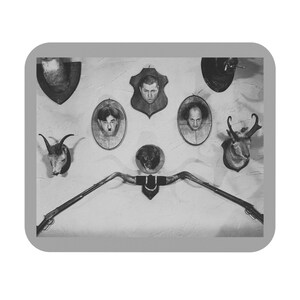 May include: Black and white image of a wall display featuring various taxidermied animal heads and framed portraits. A long, curved object resembling a rifle or horn is mounted below the portraits. The arrangement creates a vintage, eclectic aesthetic.