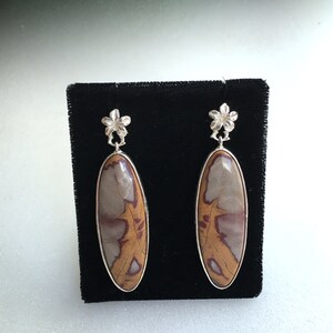 May include: A pair of silver earrings with a floral design and oval-shaped brown and white Noreena Jasper stones.
